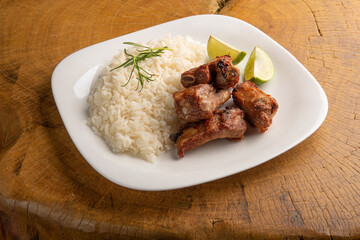 Fried pork ribs, rice and condiments on a white plate arranged on light rustic wood, selective focus.