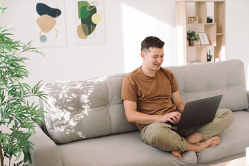 Young attractive smiling guy is browsing at his laptop, sitting at home on the cozy beige sofa at...