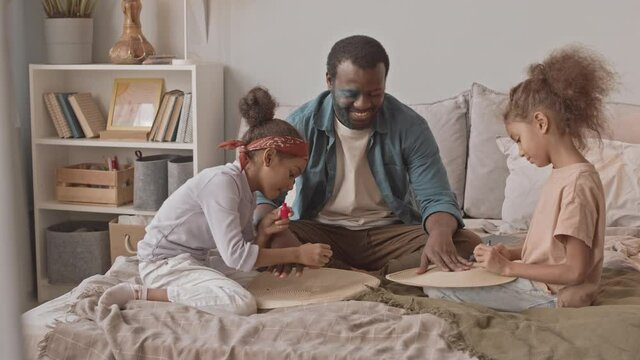 Slowmo shot of happy African-American man and his two little daughters having fun together at home. Girls painting nails of their father