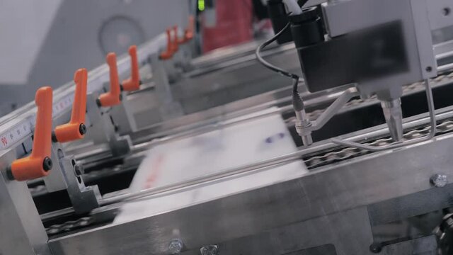 White cardboard sheet moving fast on conveyor line of automatic carton box making machine at food package exhibition, trade show - close up side view. Industrial, equipment, technology concept