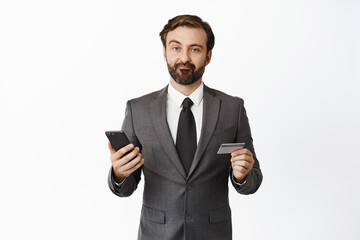 Skeptical businessman in suit grimacing disappointed, holding credit card and mobile phone, standing over white background