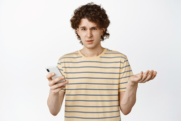 Confused young man holding phone, reacting to smth strange, staring frustrated and annoyed, standing over white background