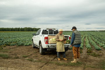 Mature Asian vegetable farmer in sunhat and apron reading papers into clipboard while talking to customer on plantation - obrazy, fototapety, plakaty
