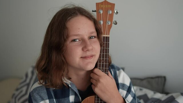  Portrait of an adorable Caucasian girl next to ukulele sitting on the bed in her room, looking at the camera smiling.