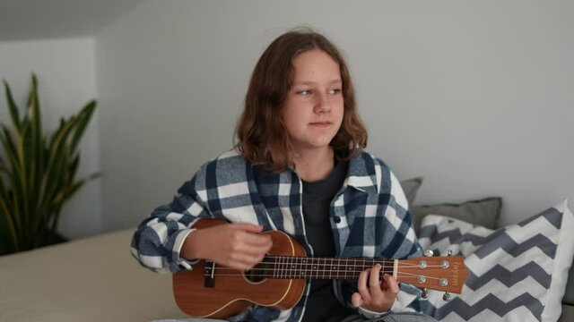 Pretty Caucasian girl playing ukulele sitting on the bed in her room enjoying happy and relaxed moments. Concept music classes and musical instruments as a hobby