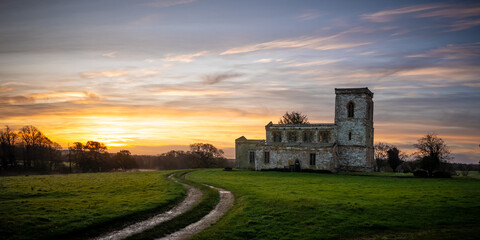 An ancient English church stands in a meadow against a backdrop of a magnificent sunrise. In the foreground a set of tractor tyre tracks leads the eye through the field.