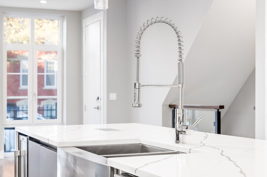 A Kitchen Sink Detail Shot With A Chrome Faucet, Stainless Steel Apron Sink, And White Granite Counter Tops Looking Out Towards A Staircase And Empty Living Area.