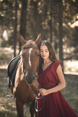 Fototapeta premium Portrait of a smiling young woman hugging her brown horse. A girl in a dress is standing near a horse. The concept of friendship between people and pets.