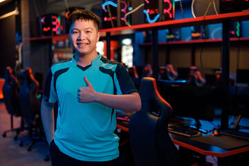 Portrait of confident Asian pro gamer happy with game results when giving thumbs-up sign in contemporary e-sports club