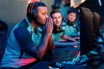 Side view of African fully concentrated esports player wearing headphones while looking at PC monitor in gaming club
