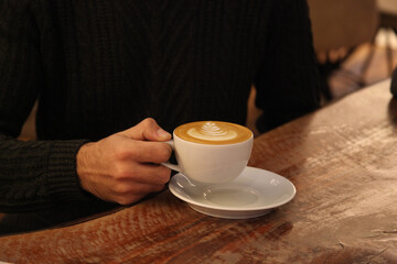 A man drinking a coffee in a cafe. He is wearing black and his face is not visible in the photo.  He is holding the coffee cup and he is sitting.