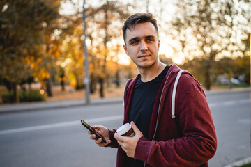 Front view of one adult young caucasian man walking in front of street in town in autumn evening holding mobile phone and cup of coffee half length real people copy space looking to the side