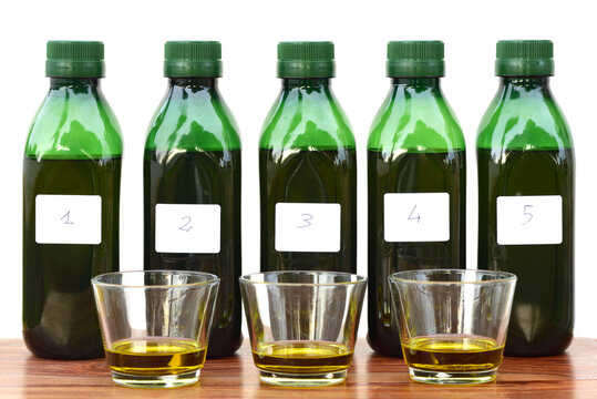 Five green plastic bottles stand next to each other and are filled with liquid. There is a number on every bottle. In front of it there are glasses filled with olive oil for testing