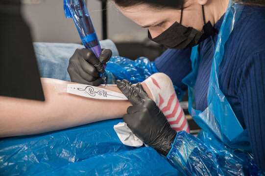 Inside a tattoo studio a young female tattoo artist works making an unalome piece on a girl's leg using a sketch.