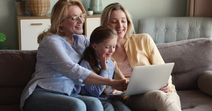 Focused little schoolgirl sit on sofa with mommy older granny engaged in funny educational activity using laptop. Adult mom middle aged grandma help small girl daughter grandchild in playing pc game