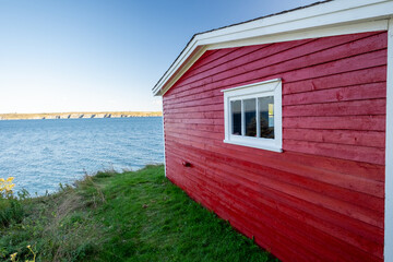 A bright red wooden building near the edge of a cliff overlooking the blue ocean. The storage building has a small glass window with white trim. In the background there's land, blue sky and blue water