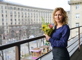 Smiling woman in a blue blouse with a bouquet of flowers stands on the balcony