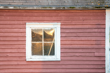 The exterior of an old red wooden vintage shed wall with a white framed closed glass single pane window. The roof is black shingles. In the window is a reflection of the sunset or evening sky.