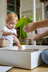 Closeup mother hand scattering sand playing at home kinetic sandbox with cute little baby