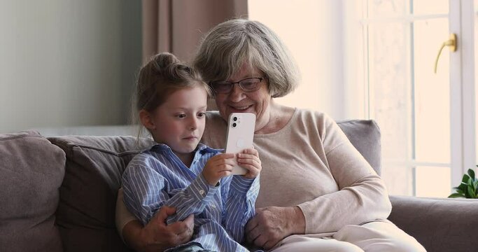 Loving old grandma sit on couch hug little grandkid watch girl playing phone game. Two diverse age female generations of one family senior granny small granddaughter discuss funny video on web channel