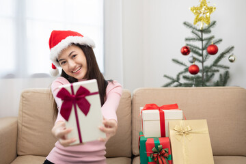 Obraz na płótnie Canvas Beautiful Asian woman showing present gift box into camera. Young Happy cute girl celebrate in christmas holiday festival giving gift box sitting on sofa at home