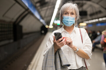 Fototapeta premium Mature European woman in mask waiting for subway train in metro station and using her smartphone.