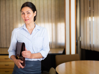 Successful confident Asian businesswoman standing near conference table in office, holding briefcase with documents..