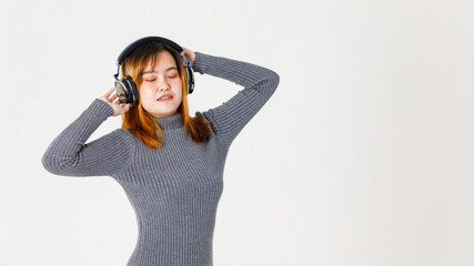 Naklejka premium Studio shot of millennial happy Asian young dyed hair female model in gray turtleneck sweater closed eyes standing listening to music via headphone headset dancing raised hands up on white background
