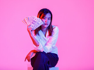 Portrait studio shot of Asian rich wealth female businesswoman model in casual wears sitting look at camera holding full hundred dollars USD cash banknote currency stack on pink light background