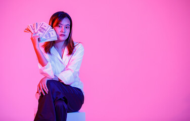 Portrait studio shot of Asian rich wealth female businesswoman model in casual wears sitting look at camera holding full hundred dollars USD cash banknote currency stack on pink light background