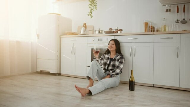 Happy home owner. Young cheerful asian woman sitting on kitchen floor with glass of wine, enjoying her own apartment