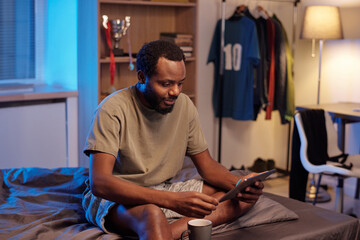 Young African man in t-shirt scrolling through news in tablet while sitting on bed after sleep in the morning