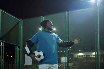 Young successful African sportsman in uniform holding soccer ball in hand while standing in front of stadium during match
