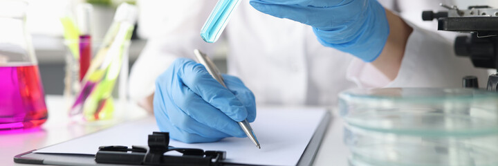 Woman scientist holds test tube with blue liquid in gloves and writes down obtained chemical data