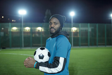 Young serious sportsman in uniform holding soccer ball while standing on football field at night