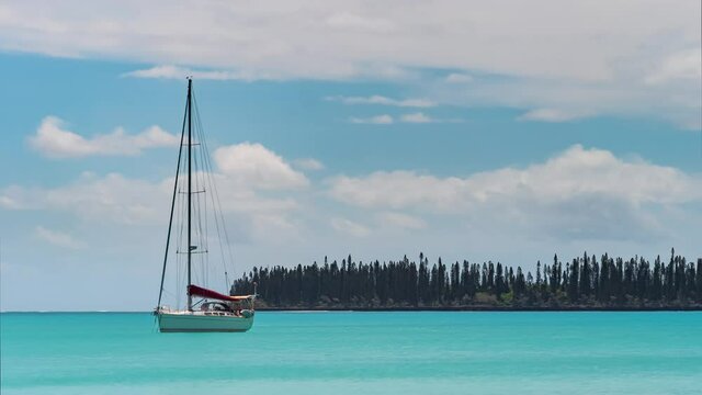 An anchored sailboat drifts in the azure blue ocean with a cloudscape overhead and Caledonian Scott pines in the background - time lapse