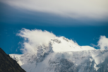 Fototapeta na wymiar Great view to high snowy mountain peaked top with low cloud under cirrus clouds in sky. Low clouds on big snow covered mountains with sharp pinnacle in sunshine. White-snow pointy peak in sunlight.
