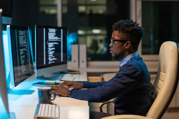 Young it-engineer of African ethnicity working with coded data while sitting in armchair in front of computer in office