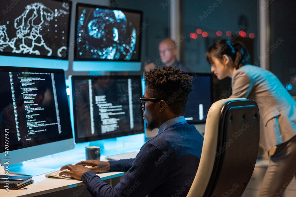 Fototapete Young it-engineer decoding data while sitting in front of computer monitors and looking at screen of one of them