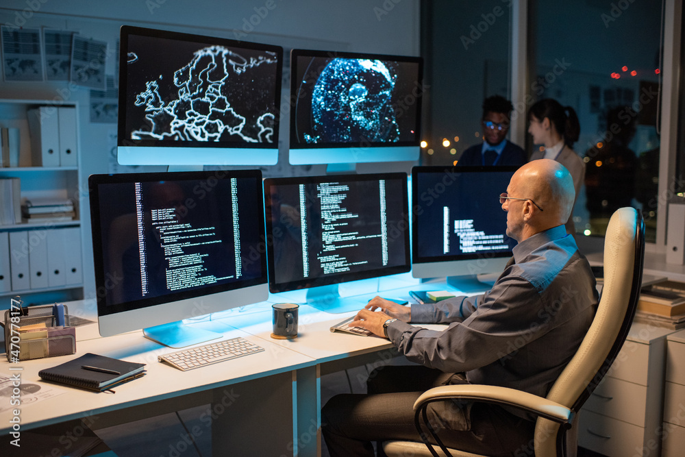 Poster Bald businessman in formalwear sitting in front of computer monitor and decoding data with two colleagues on background