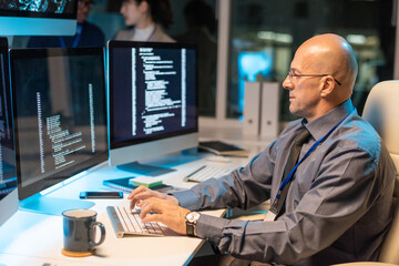 Serious elegant businessman typing and looking at coded data on computer screen while decoding it...