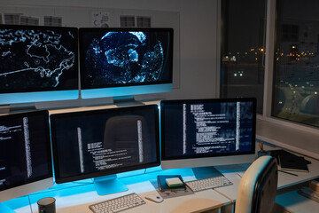 Part of large openspace office of programming company with long desk and several computer monitors with coded data