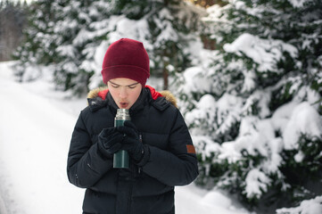 Fototapeta na wymiar Teenage boy walks through a snowy forest on a cold winter day with a hot drink in a thermos. Healthy lifestyle. Useful activity.