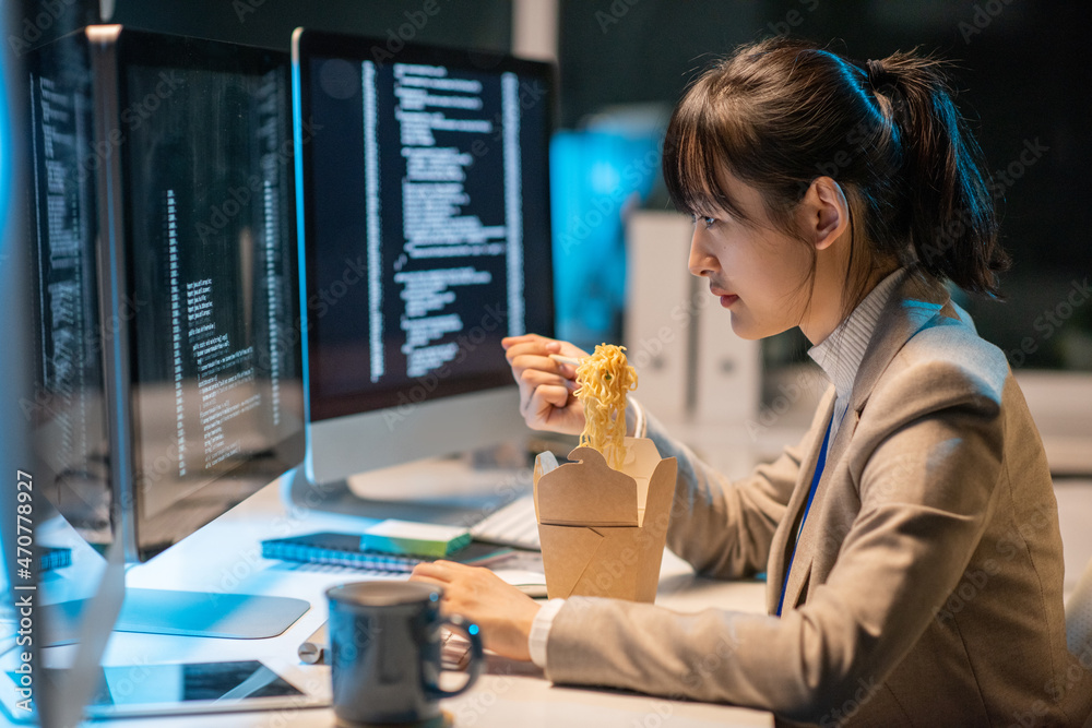 Aufkleber Side view of young Asian female programmer eating chinese wok during work over coded data in front of computer monitor