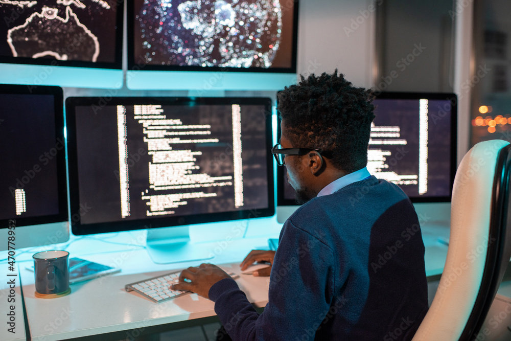 Poster Young African male software developer working late in front of computer screen with coded data in office environment