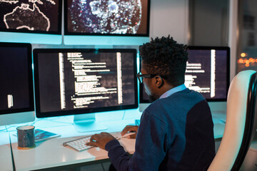 Young African male software developer working late in front of computer screen with coded data in...