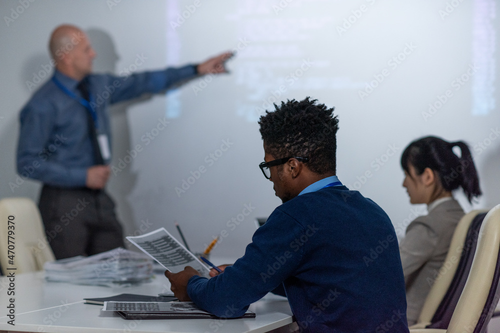 Poster young african programmer working with papers at seminar while mature businessman explaining data on 