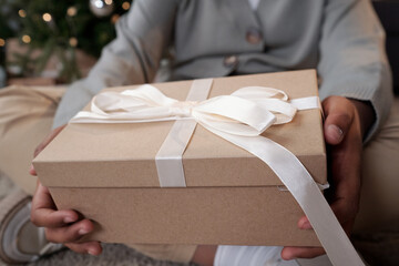 Big packed giftbox with white silk ribbon on top containing xmas present held by girl passing it to...