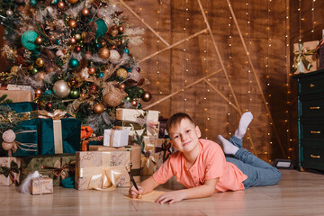 Obraz na płótnie Canvas nine-year-old boy lies on his stomach and writes on the foliage of the paper his wishes for Christmas