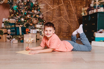 smiling eight-year-old boy lies on his stomach near a Christmas tree. letter to Santa Claus